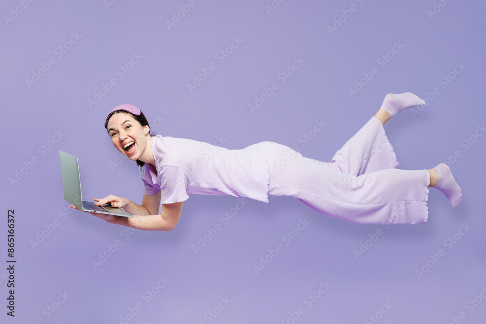 Full body sideways young IT woman in pyjamas jam sleep eye mask rest relax at home fly fall hover over air in dreams work use laptop pc computer isolated on plain purple background Night nap concept Full body sideways young IT woman in pyjamas jam sleep eye mask rest relax at home fly fall hover over air in dreams work use laptop pc computer isolated on plain purple background Night nap concept