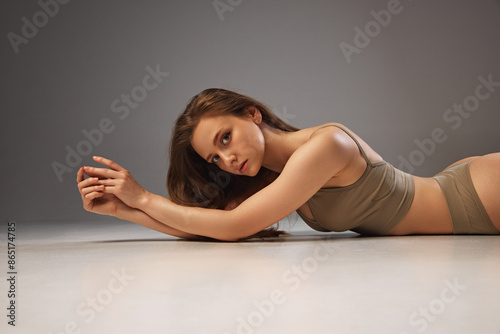 Portrait of young sensual woman poses lying on floor in beige underwear against warm grey studio background. Model with toned body. Concept of beauty and fashion, spa, bodycare, fitness, cosmetology.