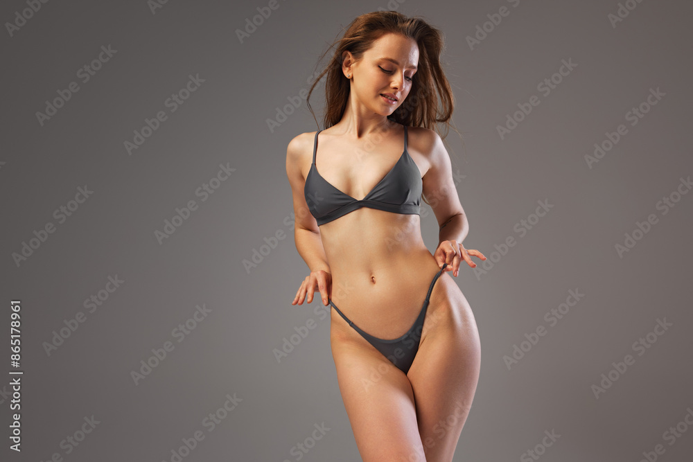 Fototapeta premium Young, beautiful, slim woman in black bikini posing showing her perfect body curves and toned skin against warm grey studio background. Concept of beauty and fashion, spa, bodycare, cosmetology.