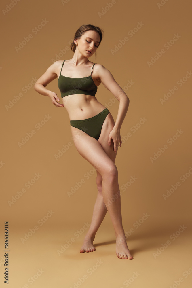 Naklejka premium Full-length portrait of young tender woman posing in khaki underwear crossed legs, shows her slim figure against sandy studio background. Concept of beauty, bodycare, anti-cellulite program, dieting.