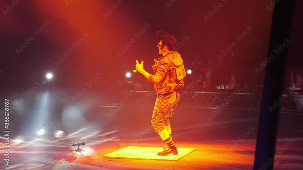 A clown with a funny hairstyle entertains guests in the circus arena in the light of colorful spotlights and smoke. He is dancing step and tapping her heels on the wooden floor. Circus performance. A clown with a funny hairstyle entertains guests in the circus arena in the light of colorful spotlights and smoke. He is dancing step and tapping her heels on the wooden floor. Circus performance.