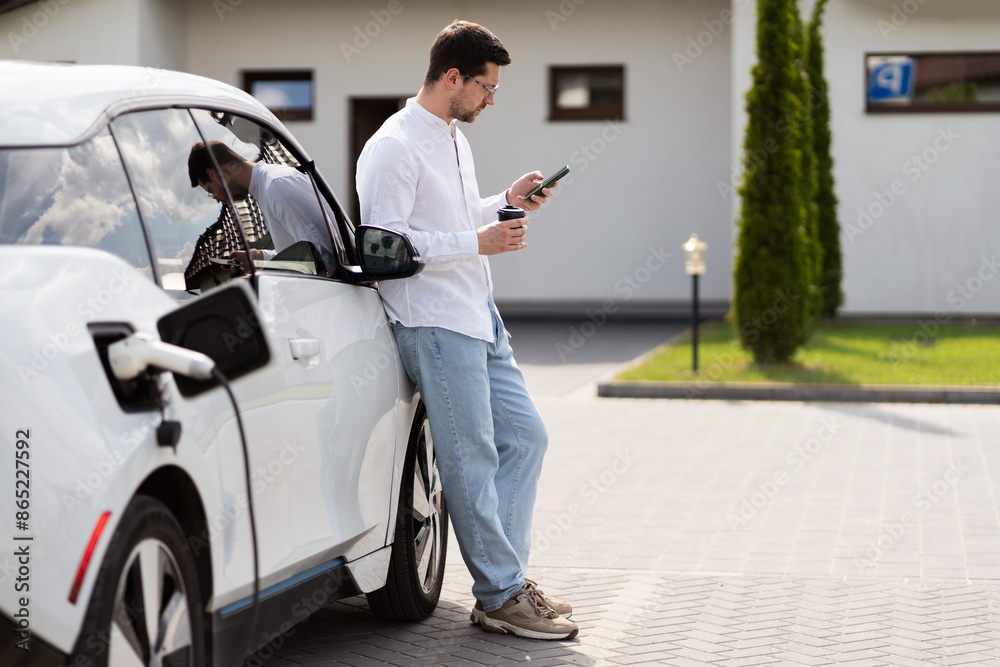 Man Charging Electric Car While Using Smartphone, Showcasing Modern Technology, Eco-Friendly Living, And Connectivity. Casual Lifestyle, Sustainable Transportation, And Everyday Urban Scene Depicted. Man Charging Electric Car While Using Smartphone, Showcasing Modern Technology, Eco-Friendly Living, And Connectivity. Casual Lifestyle, Sustainable Transportation, And Everyday Urban Scene Depicted.