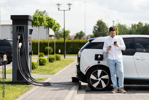 Man Charging Electric Car At Charging Station And Using Smartphone In Urban Area