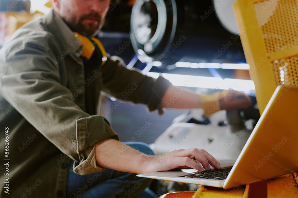 Selective focus shot of bearded repairman using laptop while fixing parts of industrial machine at work Selective focus shot of bearded repairman using laptop while fixing parts of industrial machine at work