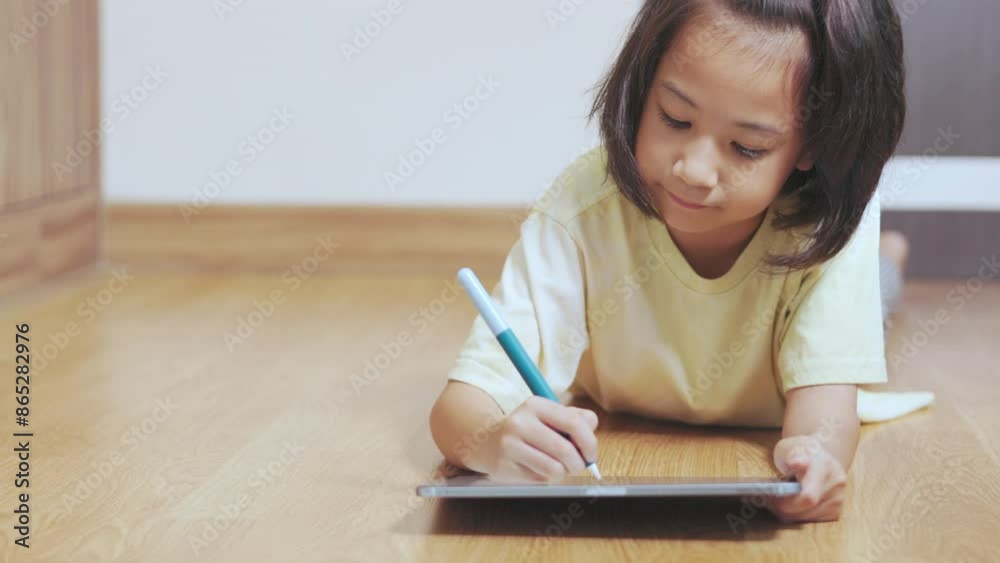 Adorable asian kid girl, drawing or writing on a tablet, lies on the floor of a cozy room. It shows the modern technology and creativity in early education of child for learning and development. Adorable asian kid girl, drawing or writing on a tablet, lies on the floor of a cozy room. It shows the modern technology and creativity in early education of child for learning and development.