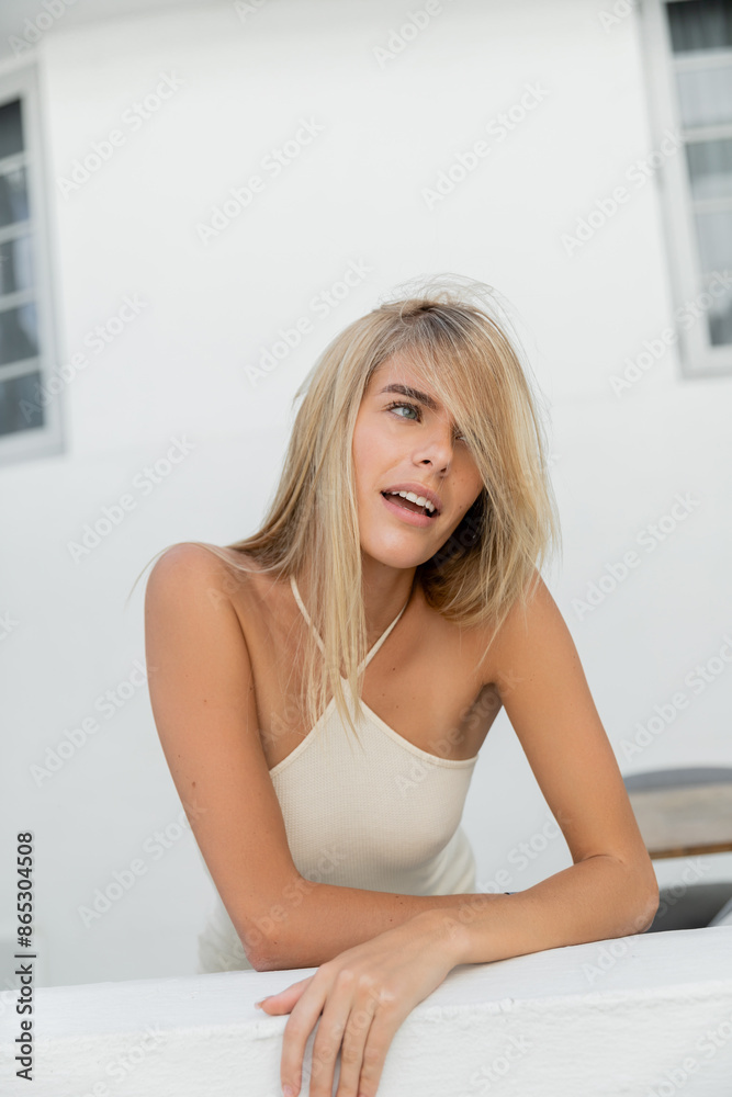 A young, blonde woman leans gracefully on a bed in a white tank top, exuding an aura of tranquility and elegance. A young, blonde woman leans gracefully on a bed in a white tank top, exuding an aura of tranquility and elegance.