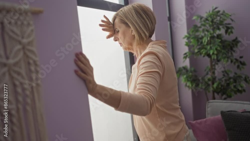 An elderly blonde woman leaning against a window in her living room, appearing dizzy in a modern apartment with purple walls and indoor plants. photo