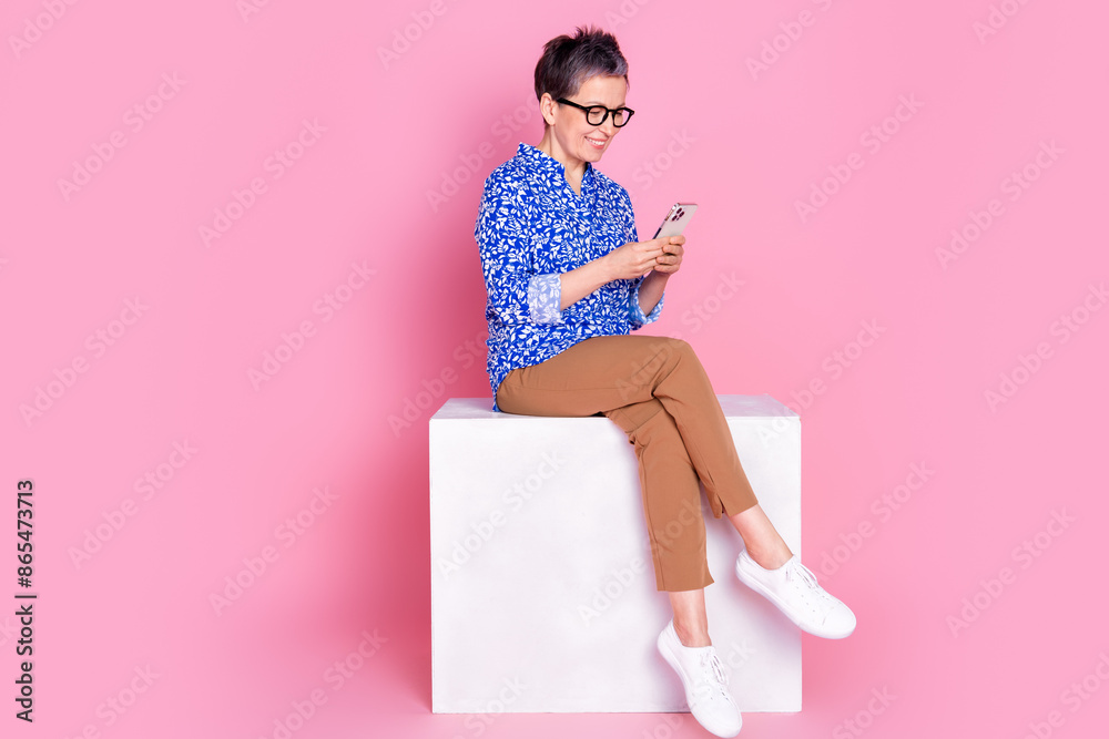 Full size photo of good mood senior person dressed print blouse sit on cube look at smartphone in hands isolated on pink color background Full size photo of good mood senior person dressed print blouse sit on cube look at smartphone in hands isolated on pink color background
