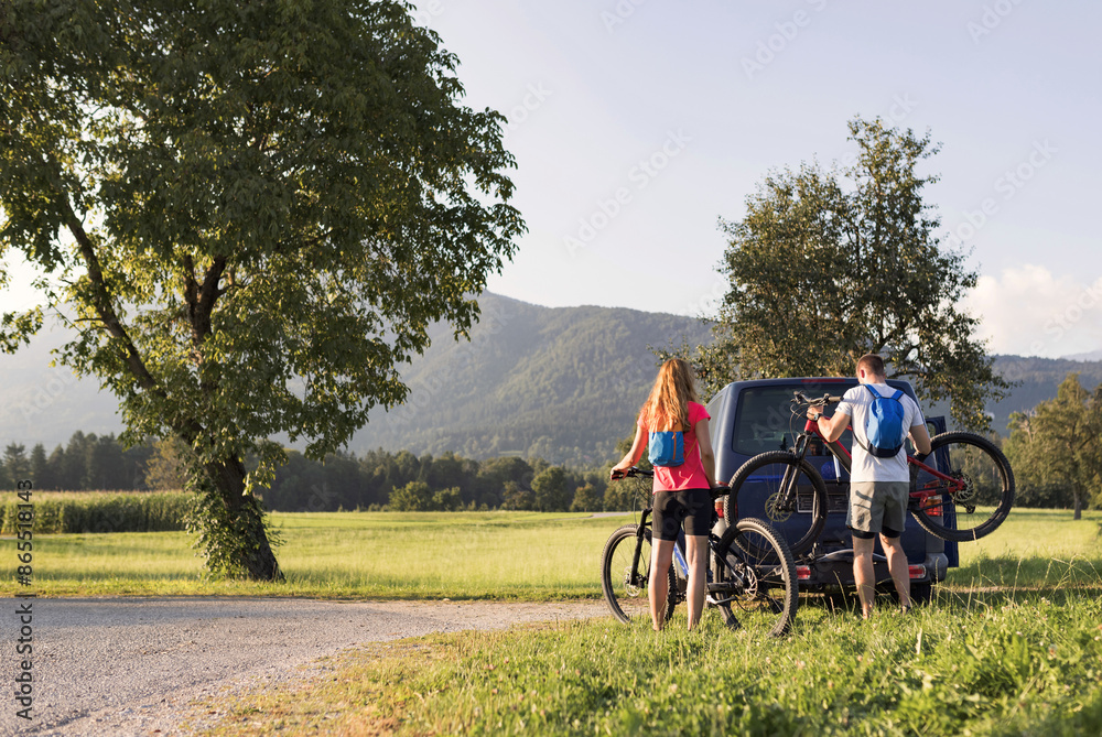 Young man is unloading electric mountain bikes, for himself and his girlfriend, lifting them from the hitch rack on the vehicle. Young man is unloading electric mountain bikes, for himself and his girlfriend, lifting them from the hitch rack on the vehicle.