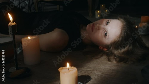 Side shot of young Caucasian lady in black dress possessed by devil waking up lying on wooden floor midst burning candles