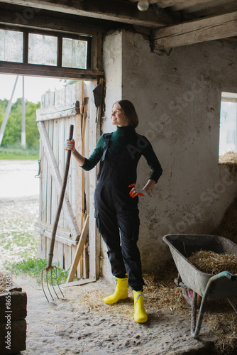 A beautiful woman farmer dressed in overalls with pitchforks in her hands is working in the cowshed. © dsheremeta