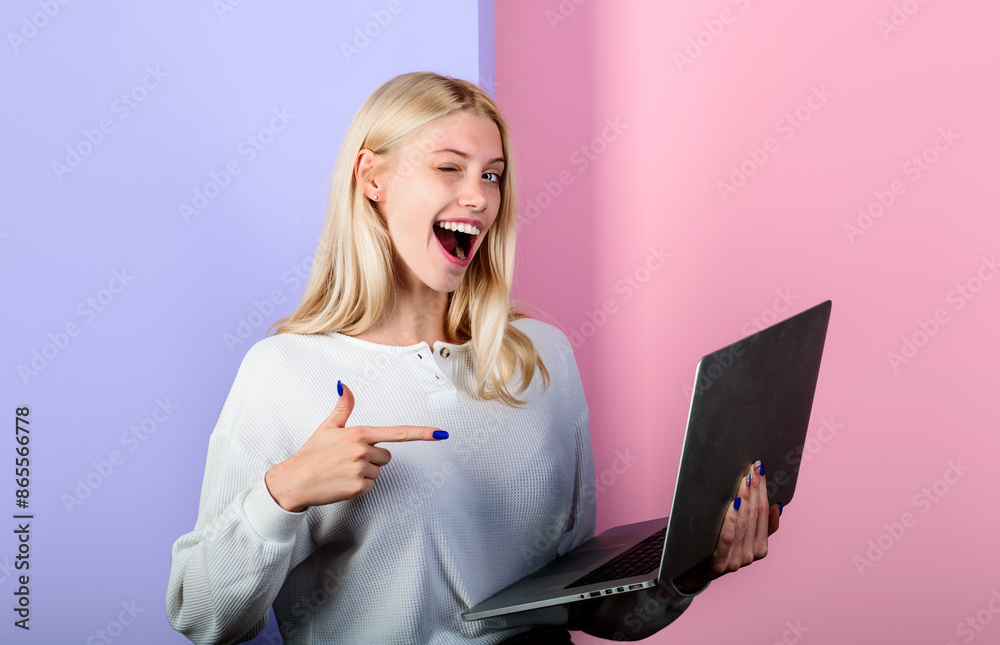 Winking girl pointing finger at laptop. Female model holding computer presenting advertising new website job search, ecommerce shopping or dating service. Smiling woman in white sweater with notebook. Winking girl pointing finger at laptop. Female model holding computer presenting advertising new website job search, ecommerce shopping or dating service. Smiling woman in white sweater with notebook.