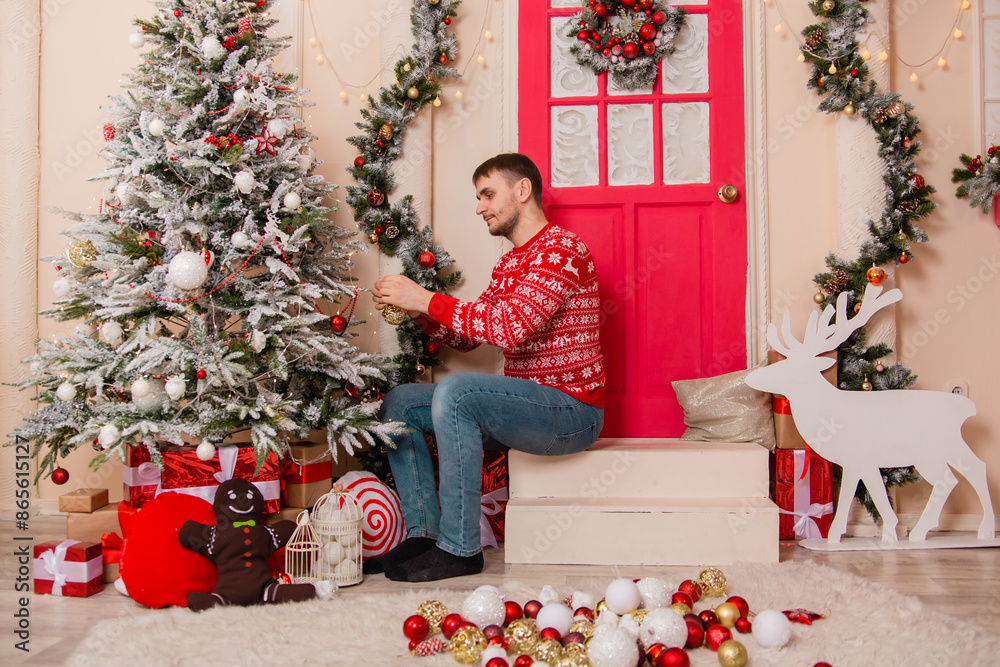 A young man in a red sweater with reindeer decorates a Christmas tree on the porch. A young man in a red sweater with reindeer decorates a Christmas tree on the porch.