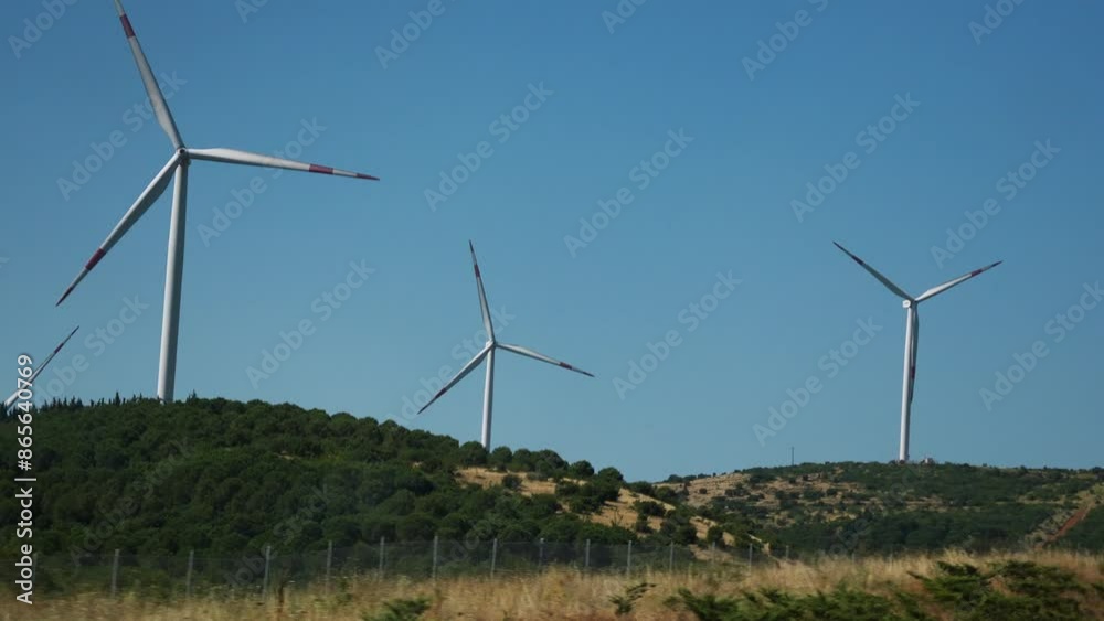 A serene landscape features large wind turbines slowly spinning on a hillside under a clear blue sky. These impressive structures are part of a renewable energy project, capturing the power of the