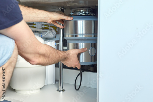 A plumber with gloved hands installs a household waste shredder for the kitchen sink. A technician installs or repairs a household waste shredder in the kitchen.