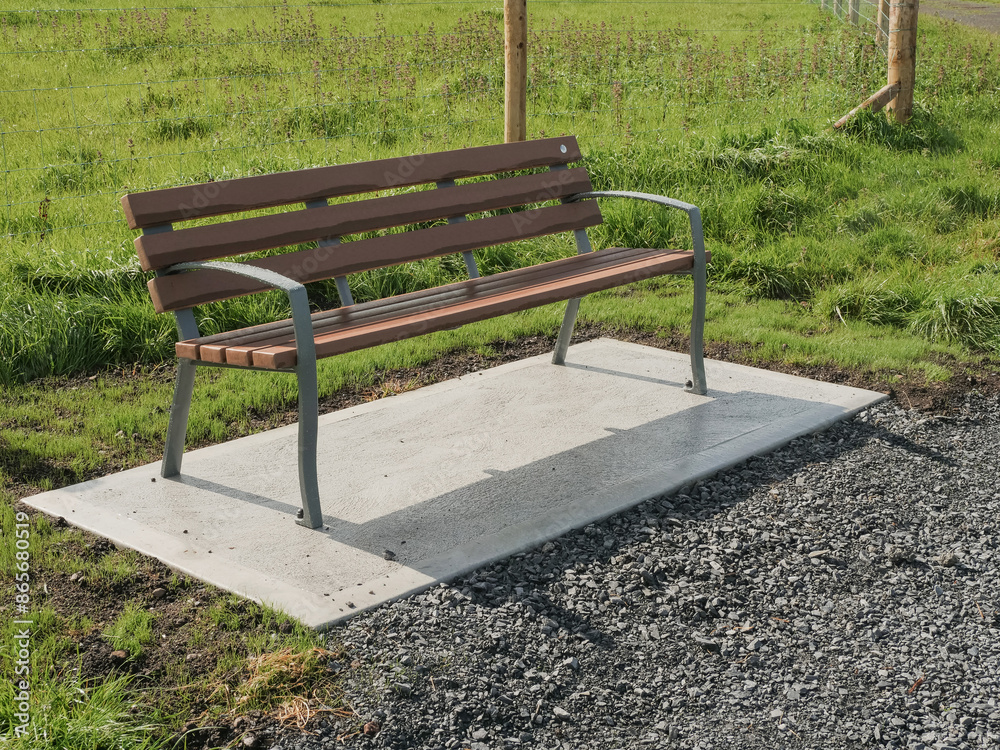 A bench is sitting on a concrete slab in a grassy area of a park. The bench is empty and the grass is green. Warm sunny day. Metal and plastic materials A bench is sitting on a concrete slab in a grassy area of a park. The bench is empty and the grass is green. Warm sunny day. Metal and plastic materials
