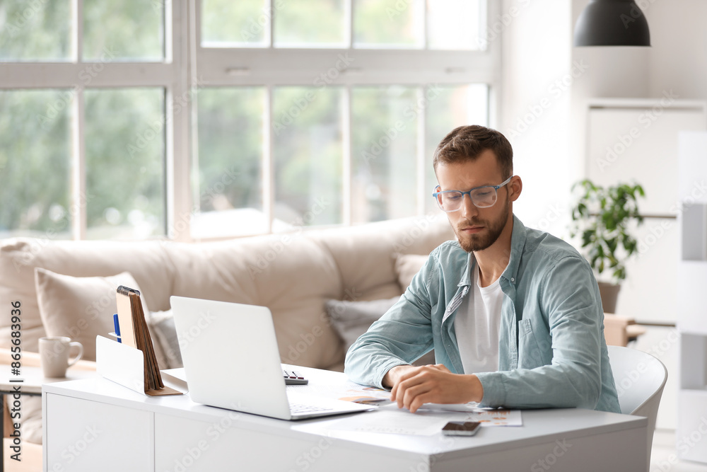 Young businessman in eyeglasses doing paper work at workplace with modern laptop in office Young businessman in eyeglasses doing paper work at workplace with modern laptop in office