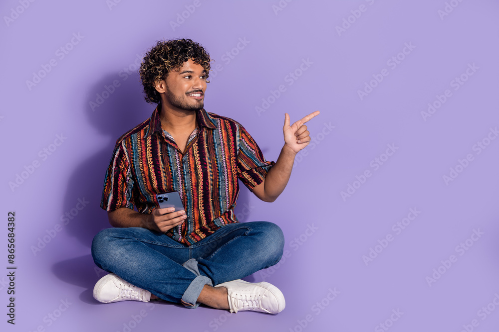 Full size photo of nice man dressed print shirt jeans sit hold smartphone look directing empty space isolated on purple color background Full size photo of nice man dressed print shirt jeans sit hold smartphone look directing empty space isolated on purple color background
