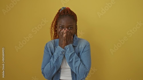 Laughing african american woman wearing denim shirt, covering mouth with hand in embarrassment, standing against isolated yellow background amidst a gossip photo