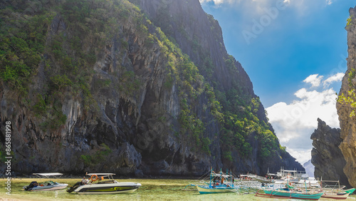 Motorboats and traditional Filipino bangkas are anchored in a turquoise lagoon. Sheer karst cliffs surround the bay. Green tropical vegetation on steep slopes. Blue sky, clouds. Philippines. Palawan. photo