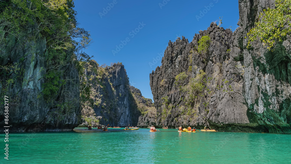 Tourists canoe in the beautiful turquoise lagoon. Sheer karst cliffs against the blue sky. Green tropical vegetation on furrowed steep slopes. Philippines. Palawan. Big lagoon. The Bacuit archipelago. Tourists canoe in the beautiful turquoise lagoon. Sheer karst cliffs against the blue sky. Green tropical vegetation on furrowed steep slopes. Philippines. Palawan. Big lagoon. The Bacuit archipelago.