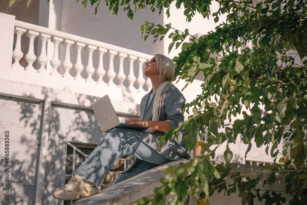 A woman is sitting on a white railing with a laptop in front of her. She is wearing a blue jacket and jeans. Concept of relaxation and leisure. A woman is sitting on a white railing with a laptop in front of her. She is wearing a blue jacket and jeans. Concept of relaxation and leisure.