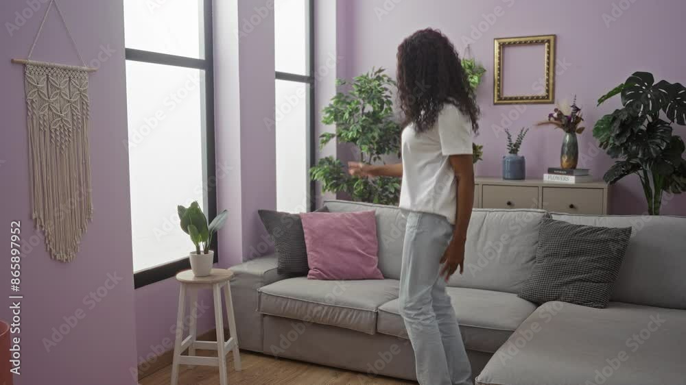 A cheerful young african american woman with curly hair is dancing in a beautifully decorated living room with purple walls, plants, and modern decor. A cheerful young african american woman with curly hair is dancing in a beautifully decorated living room with purple walls, plants, and modern decor.