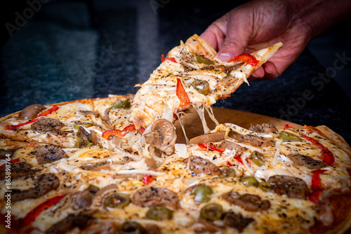 Close-up of a hand taking a slice of vegetarian pizza with onions, bell pepper, and vegetables on a wooden board.