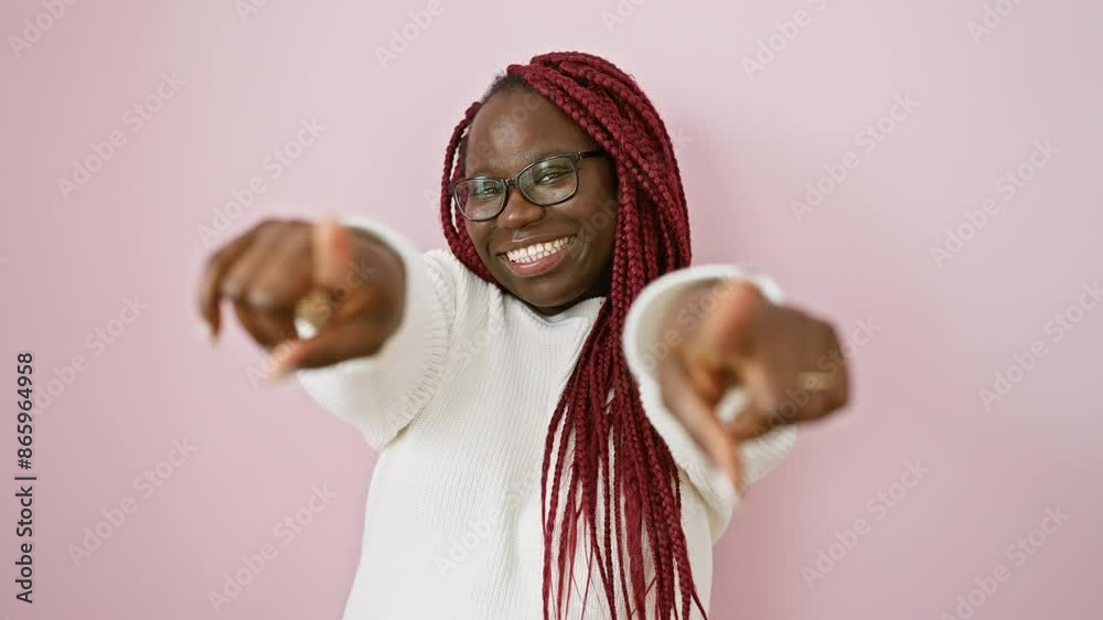 Smiling african woman with braids pointing at camera against a pink background, conveying a friendly gesture.