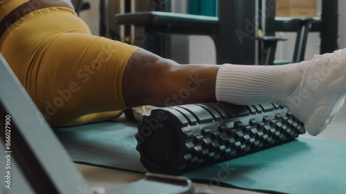 Cropped shot of unrecognizable overweight Black woman in yellow activewear using foam roller while lying on yoga mat at gym photo