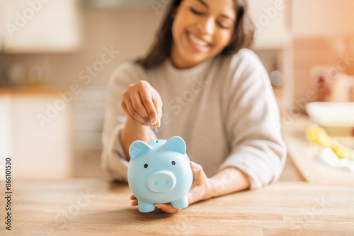 Happy Woman Putting Coin In Piggy Bank At Home Kitchen Table photo