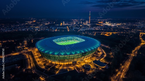 Bird's eye view of a brightly lit football stadium at night, featuring green lighting and city lights in the background. The illuminated arena is the focal point in the dark urban environment