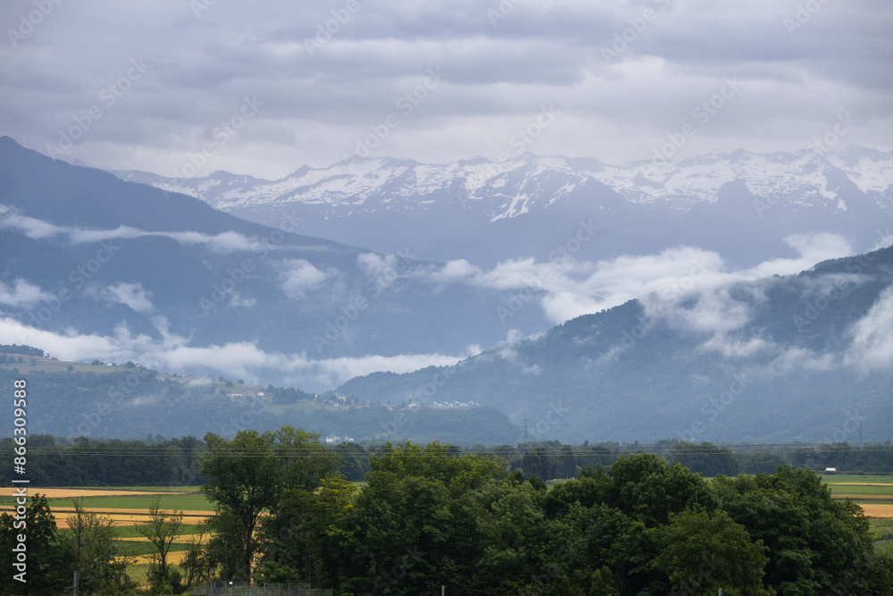 Obraz premium A scenic of mountain with layers covered with clouds, fog and snow with cloudy sky after rain in Savoie region of France . A greenery nature surrounded with French agriculture. 