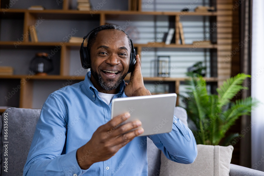 Smiling man wearing headphones enjoying music or video call on tablet in modern living room. Relaxed atmosphere with cozy decor and indoor plant Smiling man wearing headphones enjoying music or video call on tablet in modern living room. Relaxed atmosphere with cozy decor and indoor plant