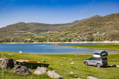 A view of Lake Turano, in the village of Castel di Tora, Rieti, Lazio, Italy. Some people do tent camping on the car, on the lake, surrounded by green mountains. Blue sky in summer.