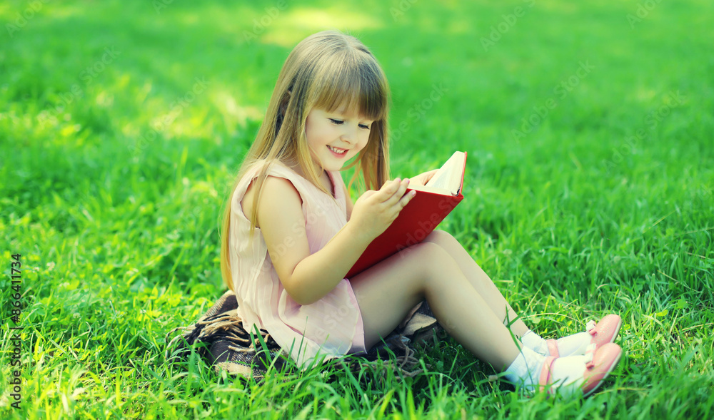 Fototapeta premium Happy little girl child reading book sitting on the grass in summer park