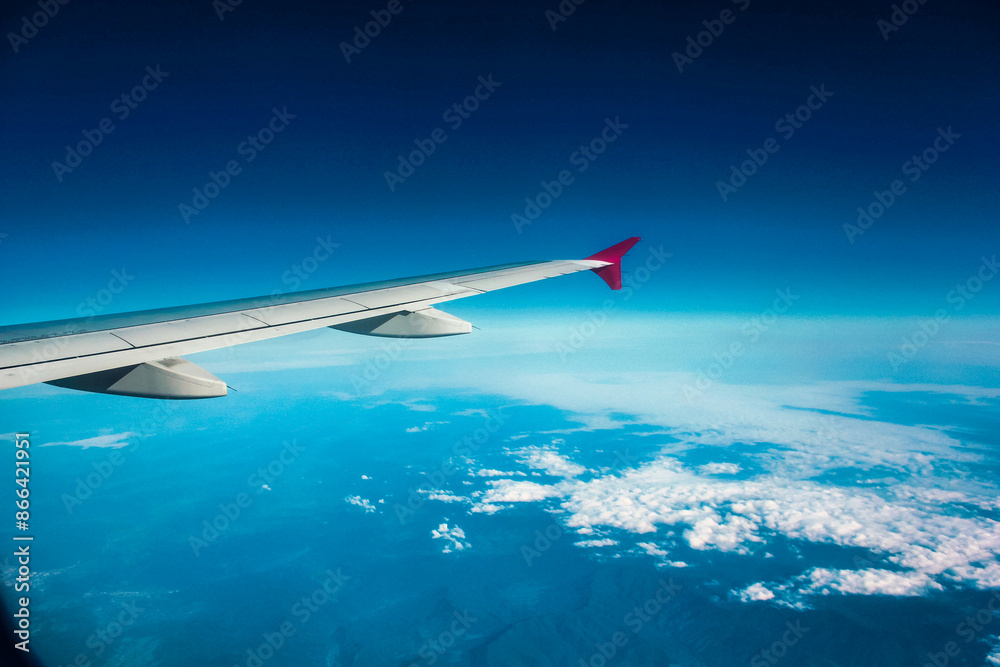 Mid-flight wing view from airplane cabin, highlighting the red winglet. Underneath, vista of land, water, and cloud-dotted sky portrays serenity in air travel. Mid-flight wing view from airplane cabin, highlighting the red winglet. Underneath, vista of land, water, and cloud-dotted sky portrays serenity in air travel.