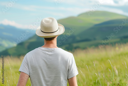 A man in a white hat and t-shirt stands amidst a lush green landscape, taking in the serene view of rolling hills.