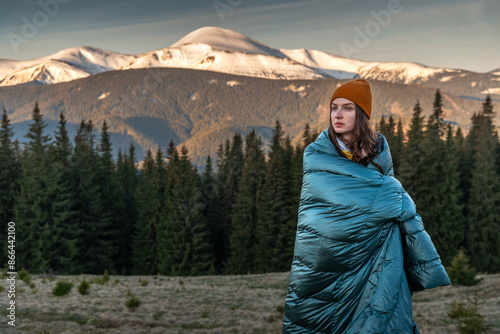 Young Woman on the Background of Snowy Mountains. Young woman wrapped in blue down blanket on the background of mountains at sunrise. Weekend adventure, people enjoy nature Carpathian Mountains photo