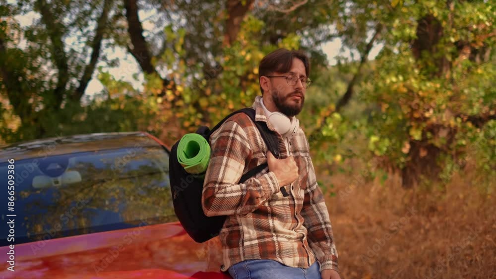 A young man sitting on the car hood with a backpack on his shoulders, enjoying warm autumn days, showcasing lightweight and spacious backpacks for hiking and road trips. A young man sitting on the car hood with a backpack on his shoulders, enjoying warm autumn days, showcasing lightweight and spacious backpacks for hiking and road trips.