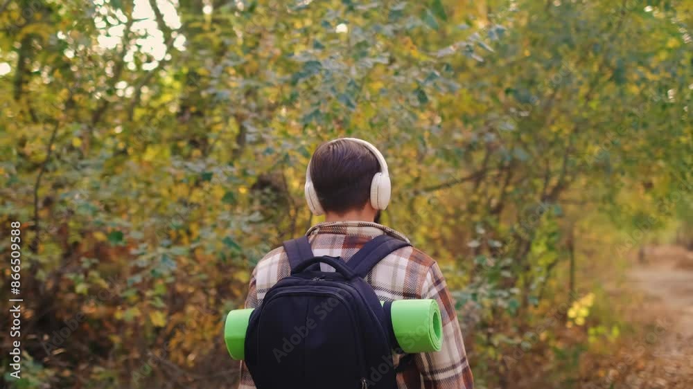 A tourist with a backpack walks away from the camera, wearing brown plaid shirt and headphones, listening to nature's music as a form of meditation. A tourist with a backpack walks away from the camera, wearing brown plaid shirt and headphones, listening to nature's music as a form of meditation.