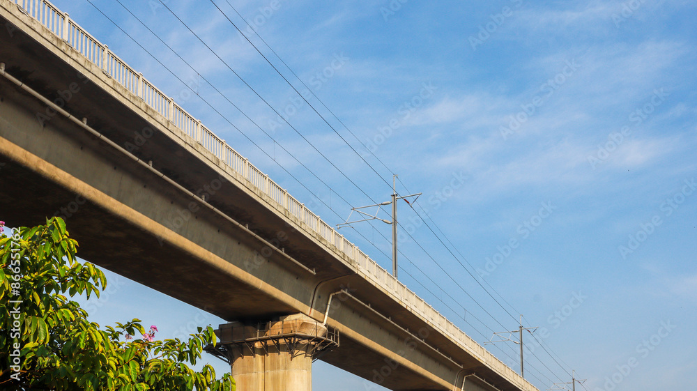 A concrete bridge with a white railing and power lines stretches across a blue sky with fluffy clouds. Elevated Concrete Bridge Against a Blue Sky A concrete bridge with a white railing and power lines stretches across a blue sky with fluffy clouds. Elevated Concrete Bridge Against a Blue Sky