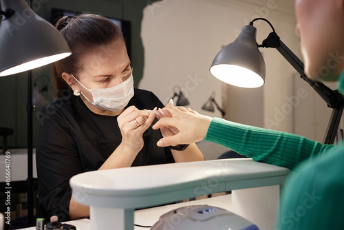 Over shoulder shot of professional female nail tech working on clients manicure wearing protective mask in well equipped modern nail salon, copy space photo
