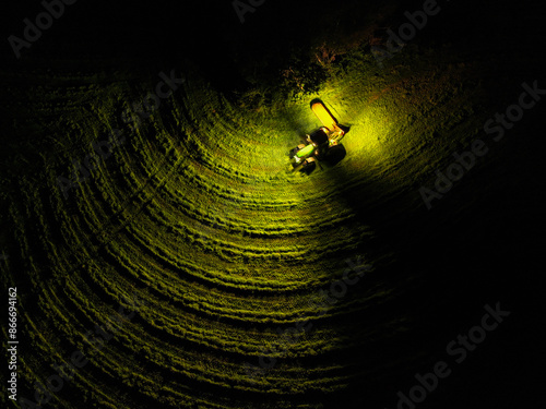 Fotografie AERIAL TOP DOWN: Tractor mowing meadow at night, illuminated by its headlights