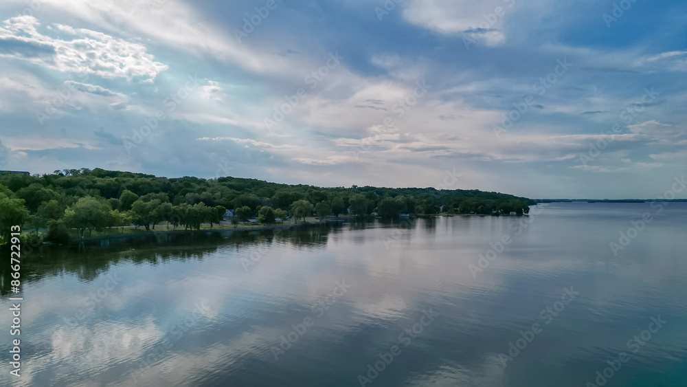 A blue sky with clouds and a shore with green trees reflected in the lake. Beautiful summer nature is mirrored in the water. Drone shot of the landscape, aerial view photo. A blue sky with clouds and a shore with green trees reflected in the lake. Beautiful summer nature is mirrored in the water. Drone shot of the landscape, aerial view photo.
