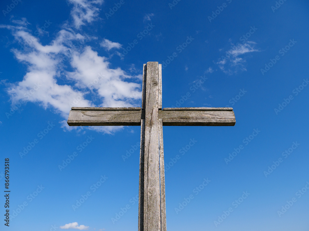 A weathered wooden cross stands tall against a bright blue sky with fluffy white clouds. A weathered wooden cross stands tall against a bright blue sky with fluffy white clouds.