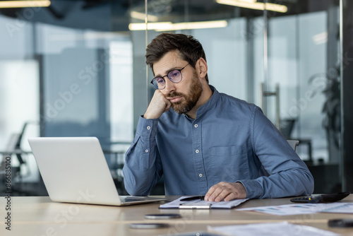 Businessman feeling bored and unmotivated working at laptop in modern office setting