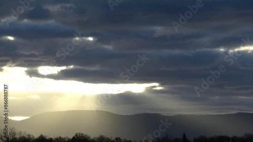 Cloudy sky with sunlight, yellow rays and dark clouds after sunrise over dark horizon with trees and Silesian Beskid mountains in Poland in December - real time. Topics: sun energy, sunrise, dawn