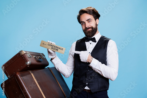 Skilled bellhop has conference room sign in hand, pointing at wall indicator to lead hotel guests to all locations on the premises. Stylish doorman posing with a pointer symbol.