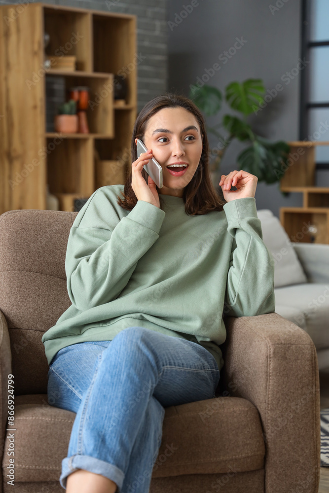 Obraz premium Happy young woman talking by mobile phone in armchair at home